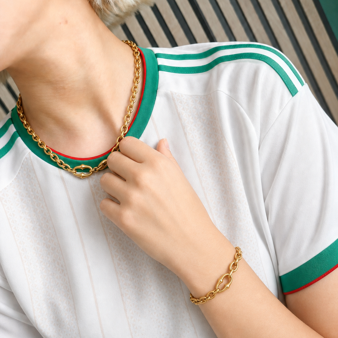 A close-up lifestyle shot of a model wearing a gold chain necklace with a green and red woven trim detail, matching gold chain earrings, and a gold chain bracelet, styled with a white Gucci t-shirt.