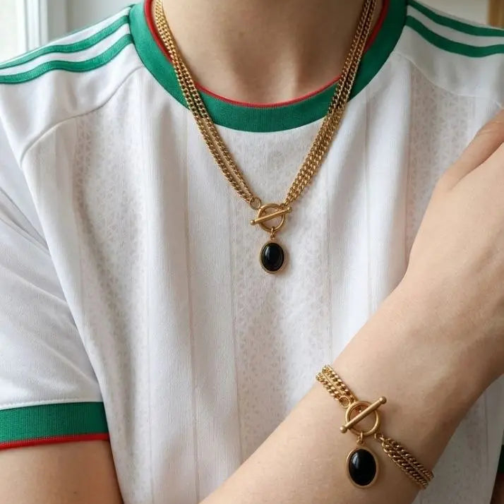 A close-up lifestyle shot of a person wearing a gold chain necklace and matching bracelet with black oval charms, styled with a white athletic t-shirt featuring green and red trim.
