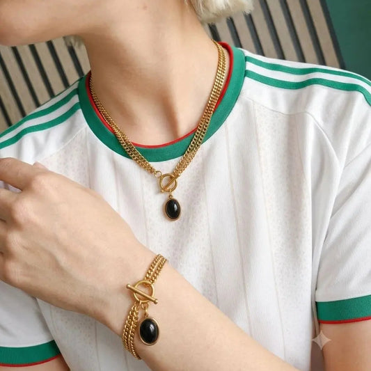A close-up lifestyle shot of a model wearing a gold chain necklace with a black teardrop pendant, a matching gold bracelet, and a gold flower-shaped earring, paired with a white and green striped athletic t-shirt.