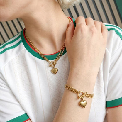 A close-up lifestyle shot of a person wearing gold jewelry, including a heart-shaped pendant necklace, a matching heart charm bracelet, and a white flower earring, styled with a white Gucci jersey featuring green and red stripes.