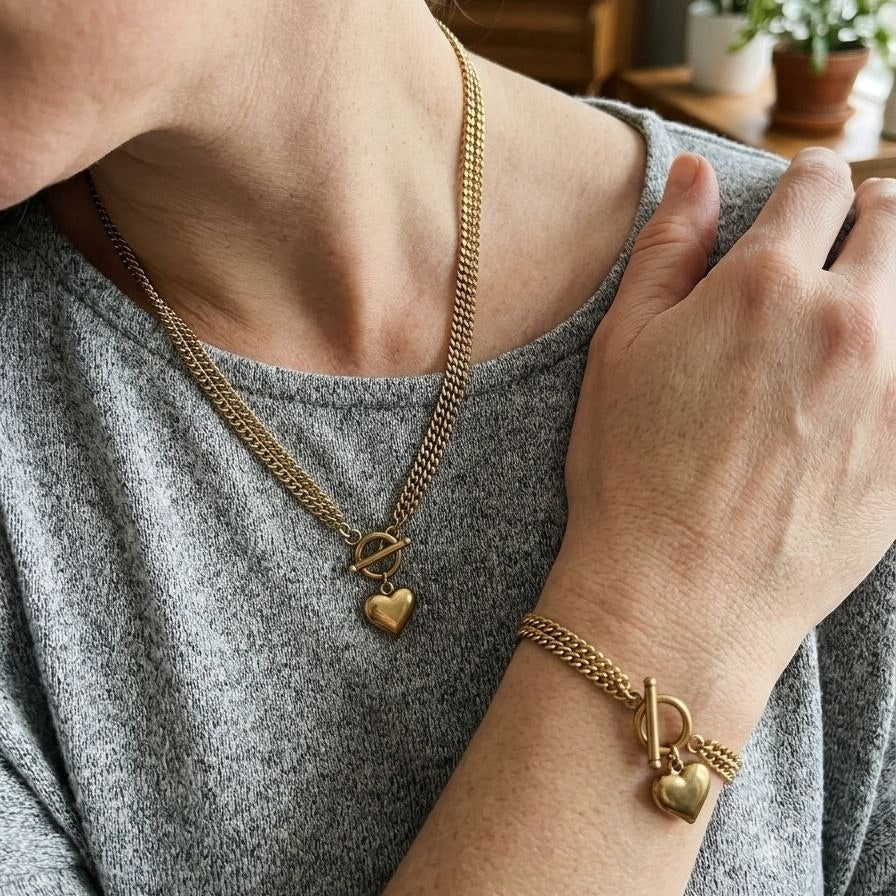 A close-up lifestyle shot of a person wearing a gold chain necklace with a heart charm and a matching gold bracelet with a heart charm, styled with a gray heathered top.